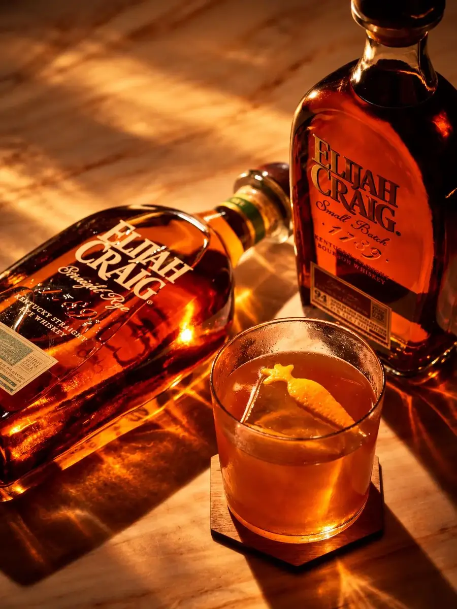Two bottles of Elijah Craig and an Old Fashioned garnished with orange peel on a wooden bar in warm amber light
